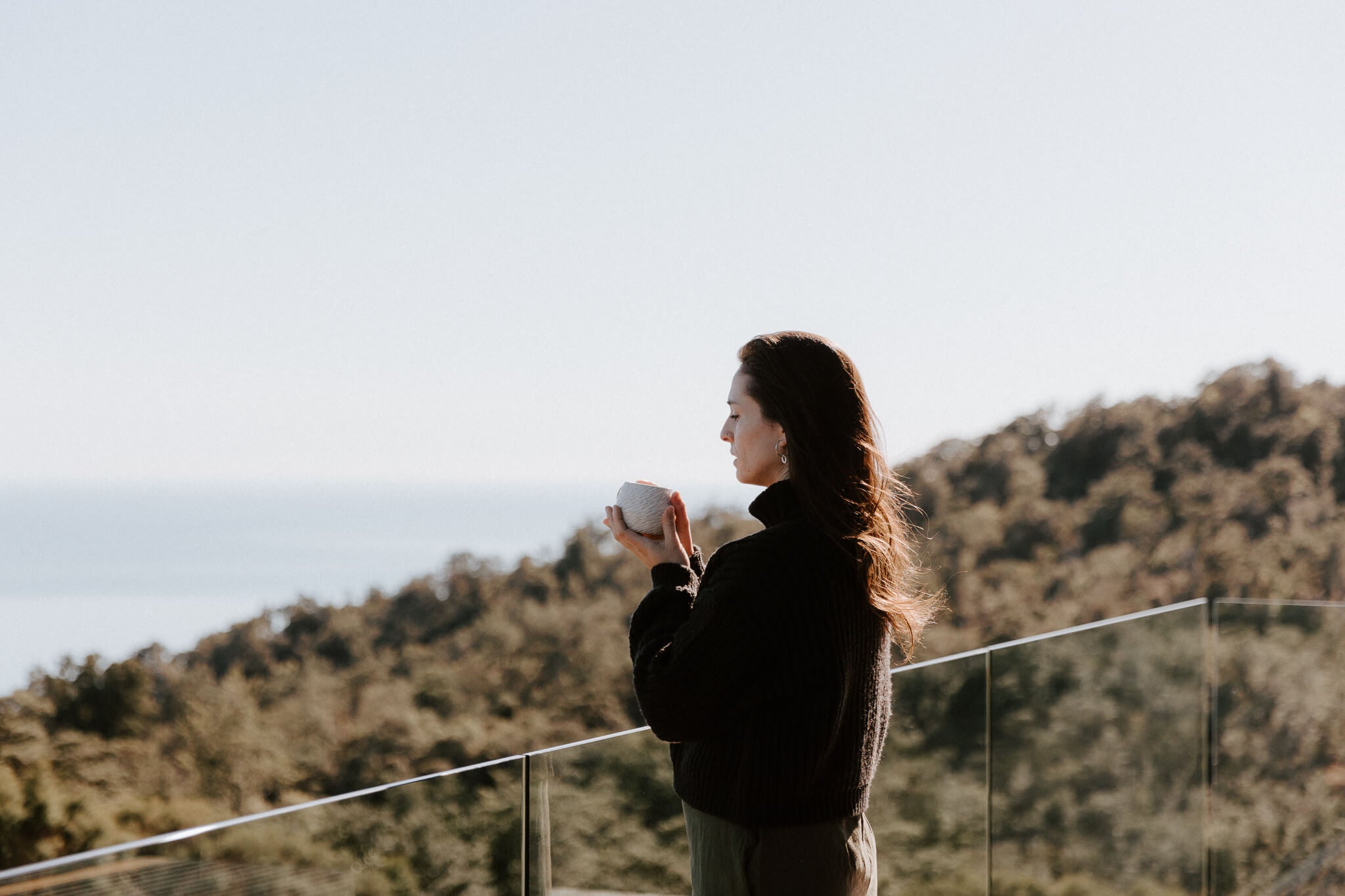 Woman holding a mug, standing on a balcony with a scenic view of trees and sky in Nelson New Zealand.