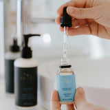 A person holding a dropper bottle of Hyaluronic Serum over a bathroom sink, with skincare products in the background.