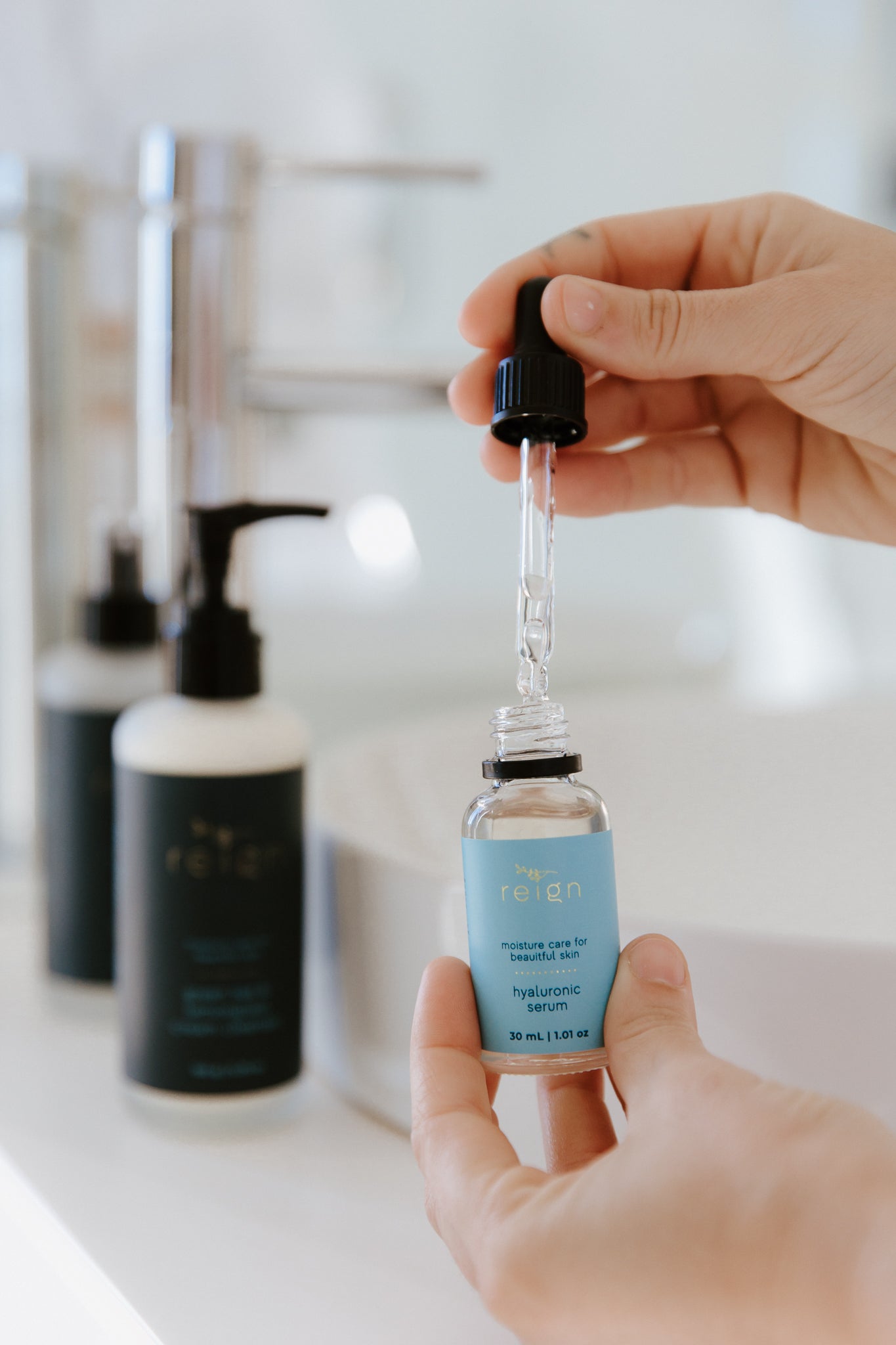 A person holding a dropper bottle of Hyaluronic Serum over a bathroom sink, with skincare products in the background.