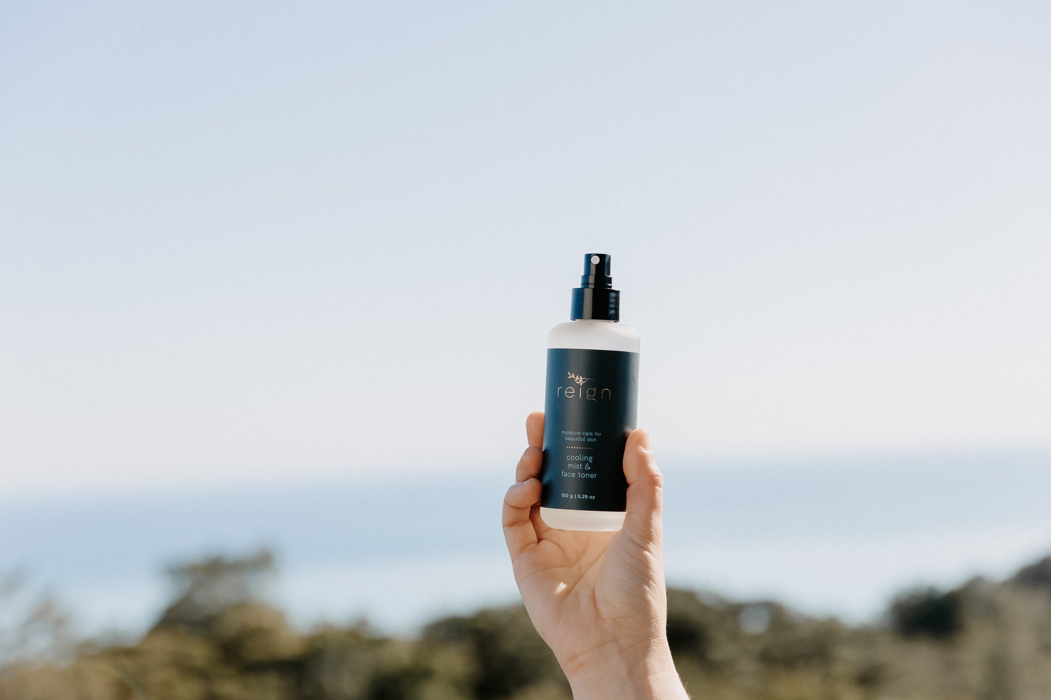 Hand holding a bottle of Reign Cooling Mist and Face Toner on a blurred natural background