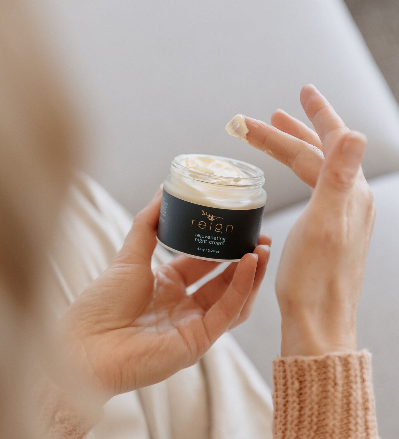 Person holding a jar of Reign Rejuvenating Night Cream product with a blurred background in a cosy setting.