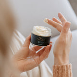 Person holding a jar of Reign Rejuvenating Night Cream product with a blurred background in a cosy setting.