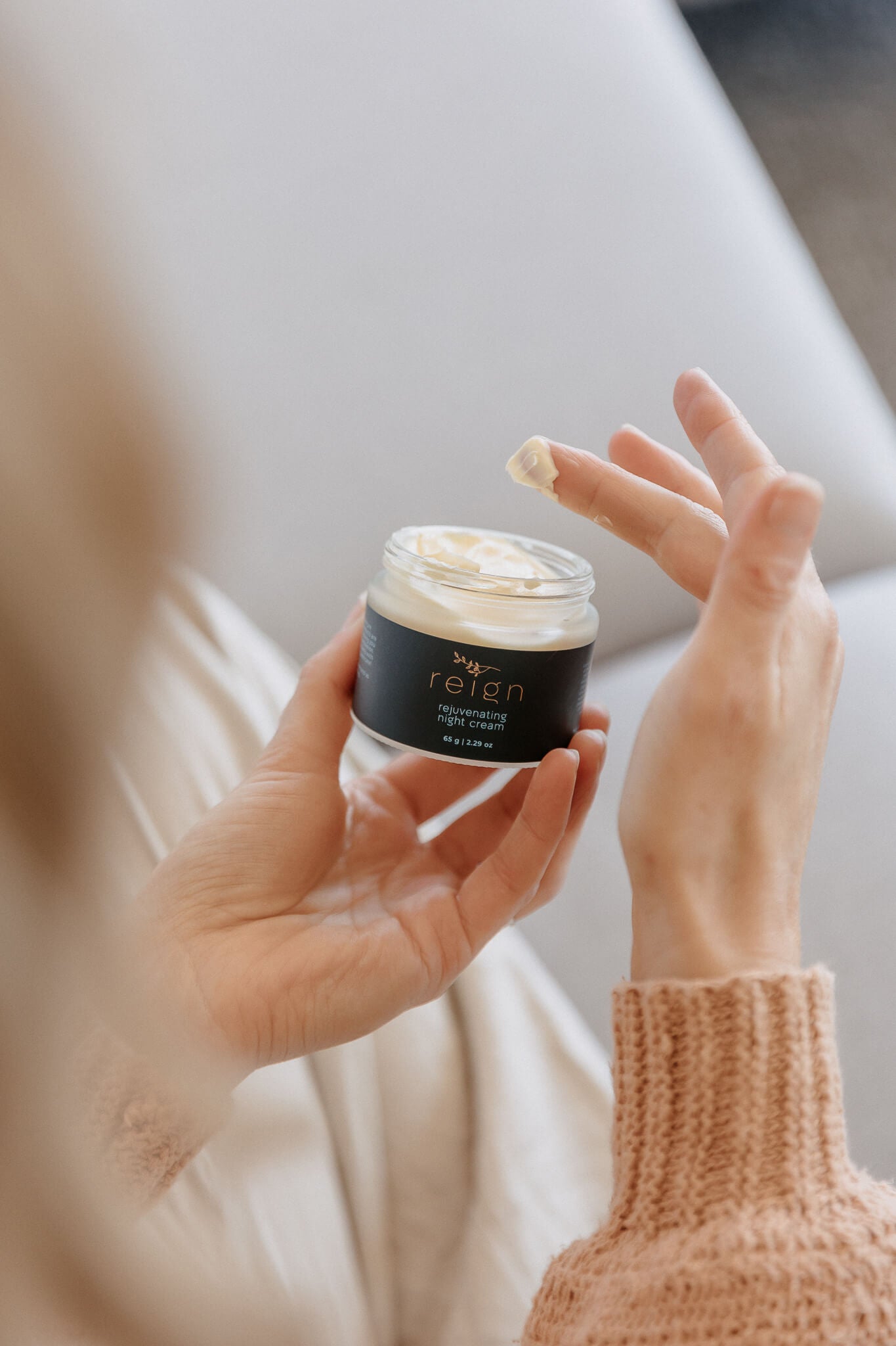 Person holding a jar of Reign Rejuvenating Night Cream product with a blurred background in a cosy setting.