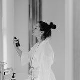 A woman spraying Reign Cooling Face Mist and Toner onto her face in a bathroom