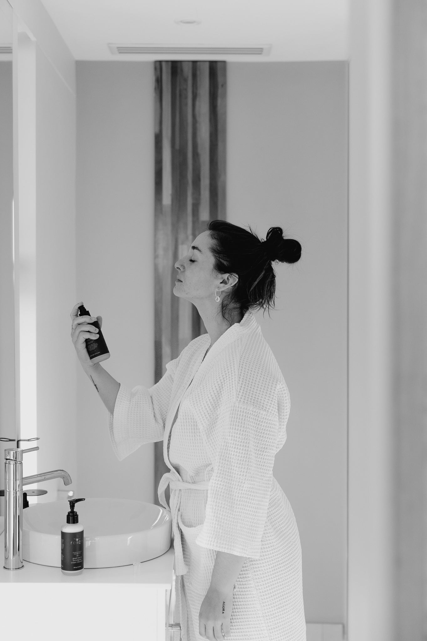A woman spraying Reign Cooling Face Mist and Toner onto her face in a bathroom