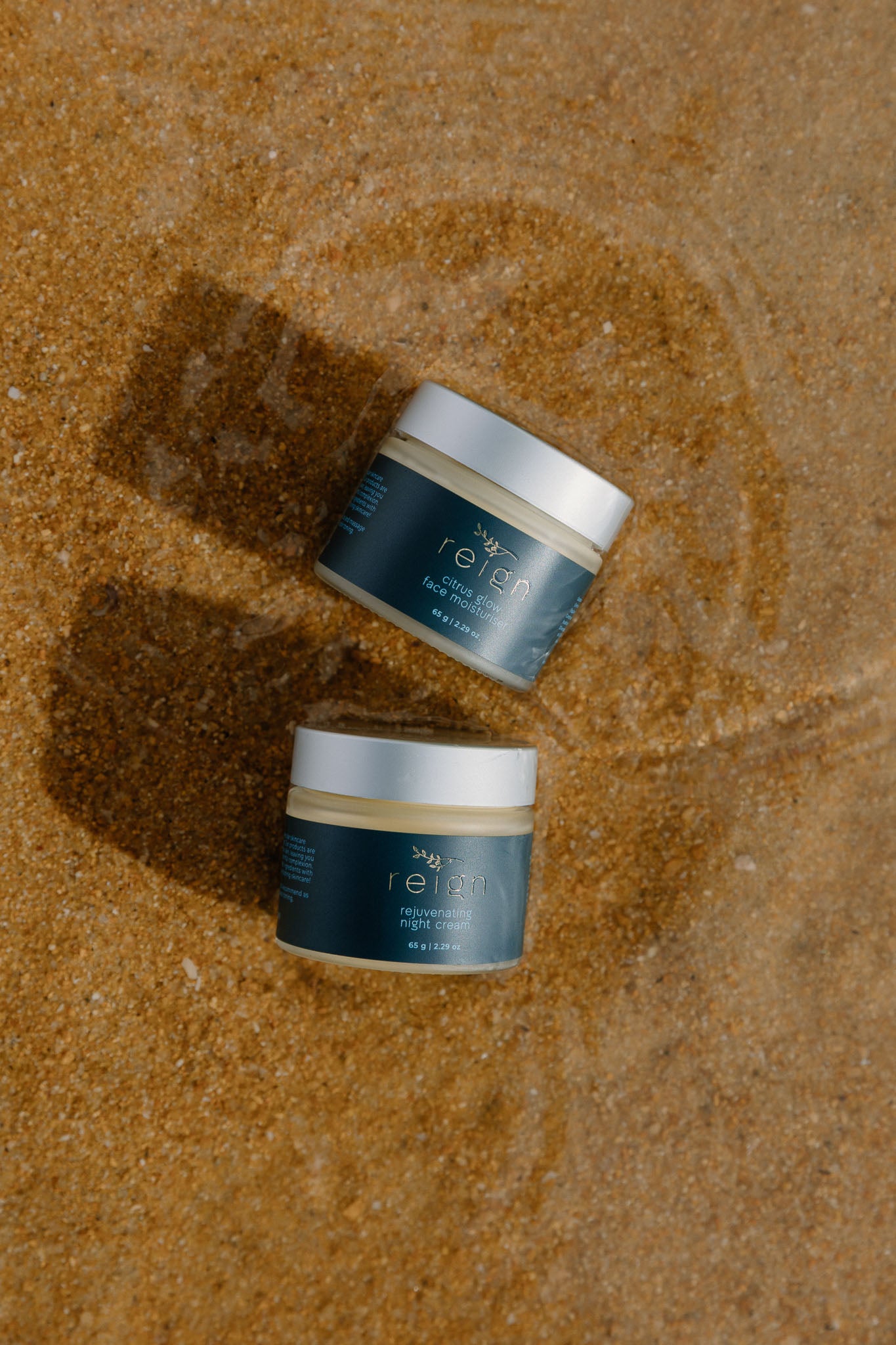 Two jars of Reign Moisturiser product on a sandy Nelson beach